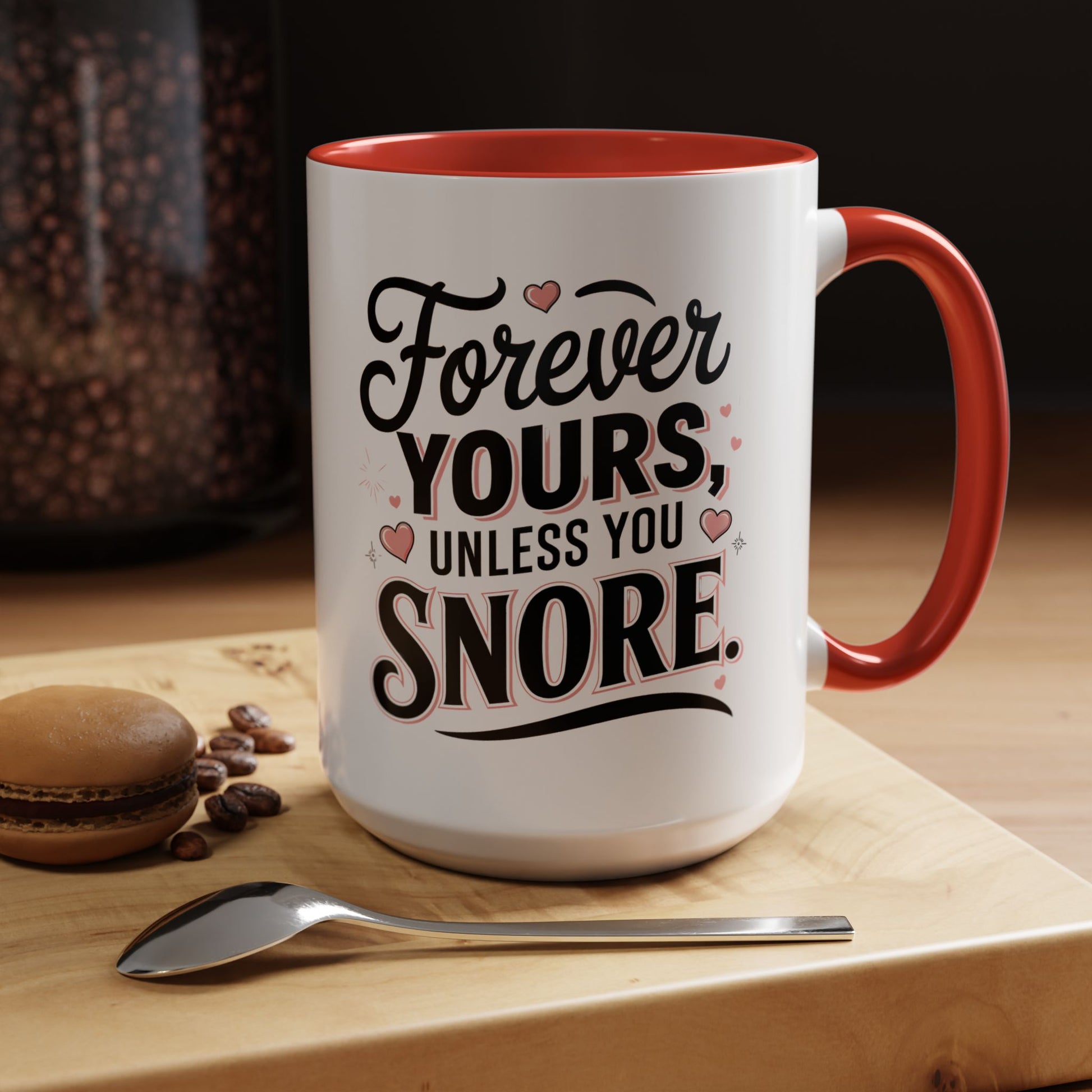 White mug with red interior and handle, featuring a humorous message, on a wooden surface with coffee beans and a spoon.